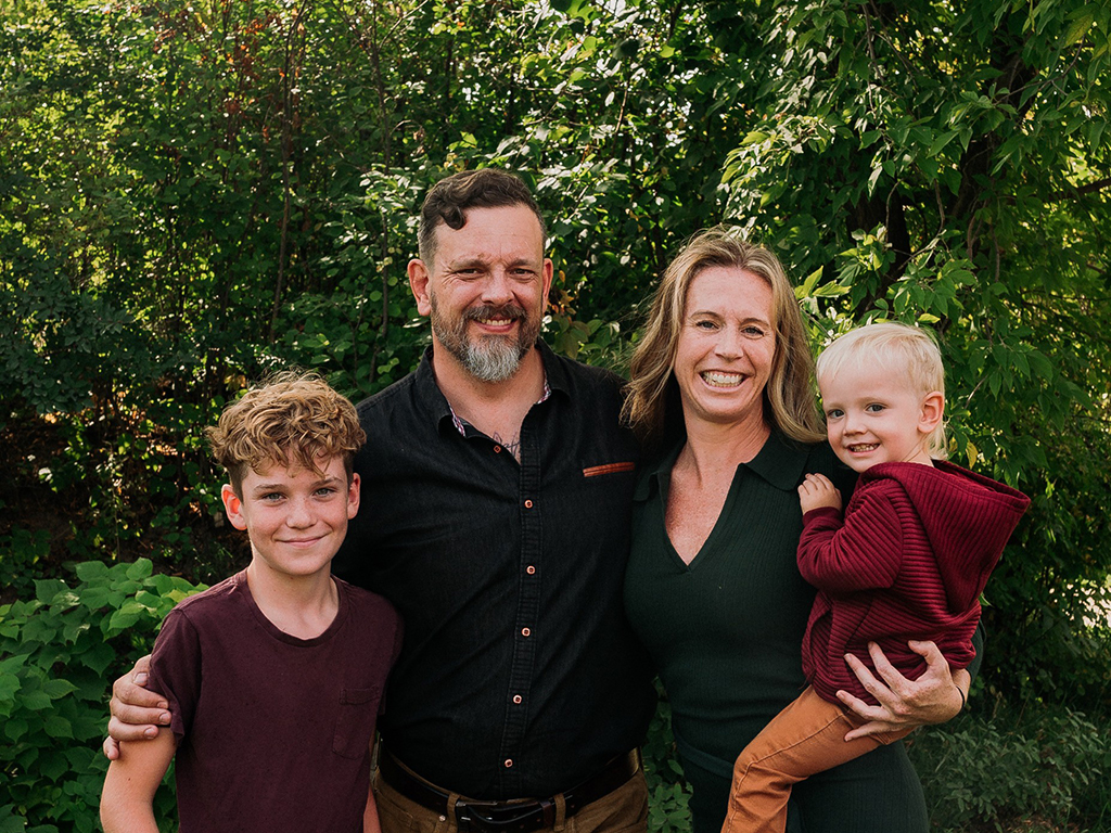 Sean, Erin and their children, smiling outdoors, surrounded by green trees.