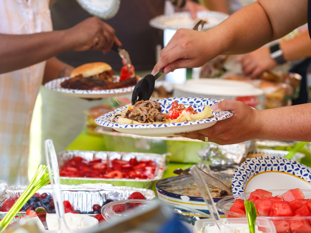 potluck dinner spread with people loading food onto paper plates